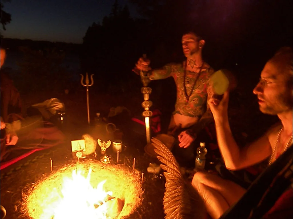 Three people sitting on ground around a cleft in the ground with small fire in the center, ritual objects around the dhuni.