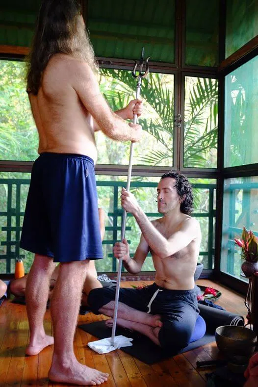 Kapilnath standing and holding a trishul, yogi seated holding onto trishul, tropical setting.