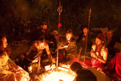 8 people sitting on ground, at night, in front of a cleft in the ground, illuminated with fire, trishuls